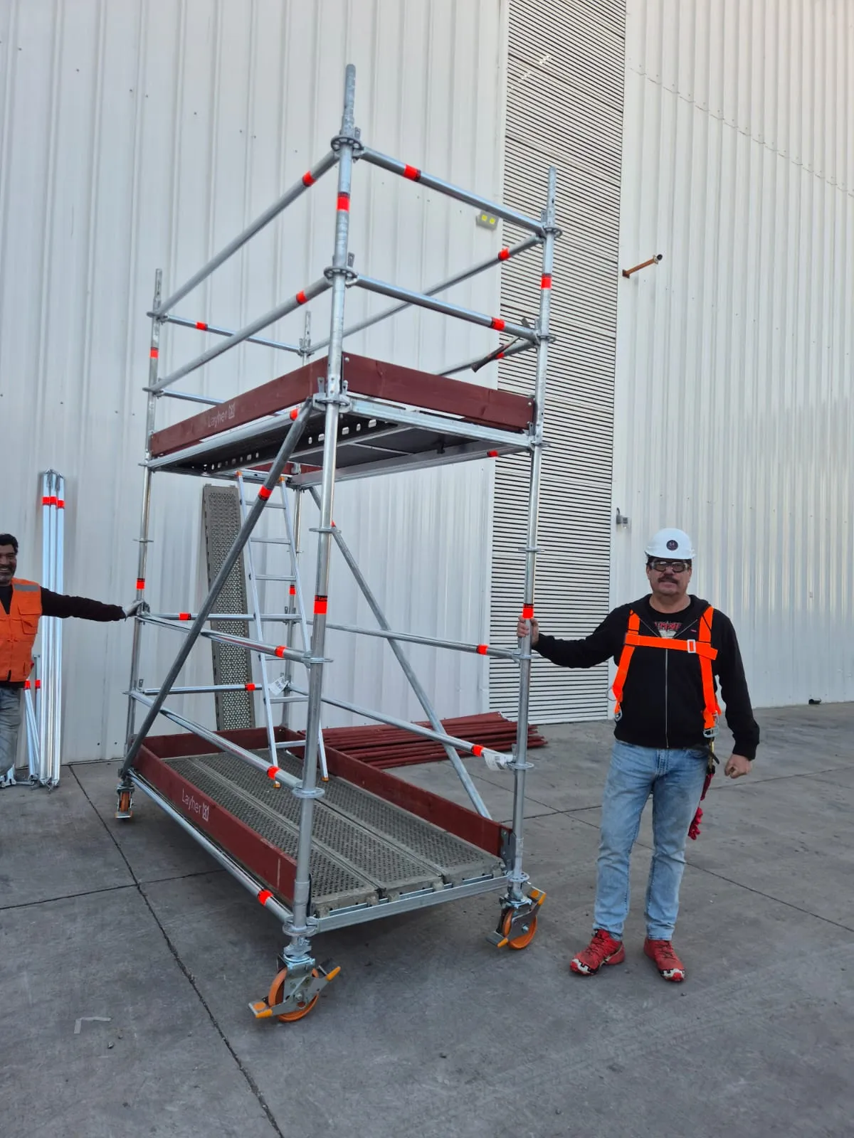 Torre móvil Layher de dos niveles con ruedas naranjas y operario con equipo de seguridad junto a la estructura
